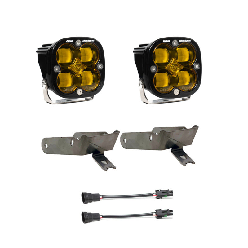 The image shows a Baja Designs 1999-2010 Ford Super Duty Fog Pocket Kit SAE Amber, featuring off-road lights with amber lenses, two black mounting brackets, and two wiring harnesses. The square-shaped lights have rounded edges with four bulbs each, ideal for rugged terrain adventures.